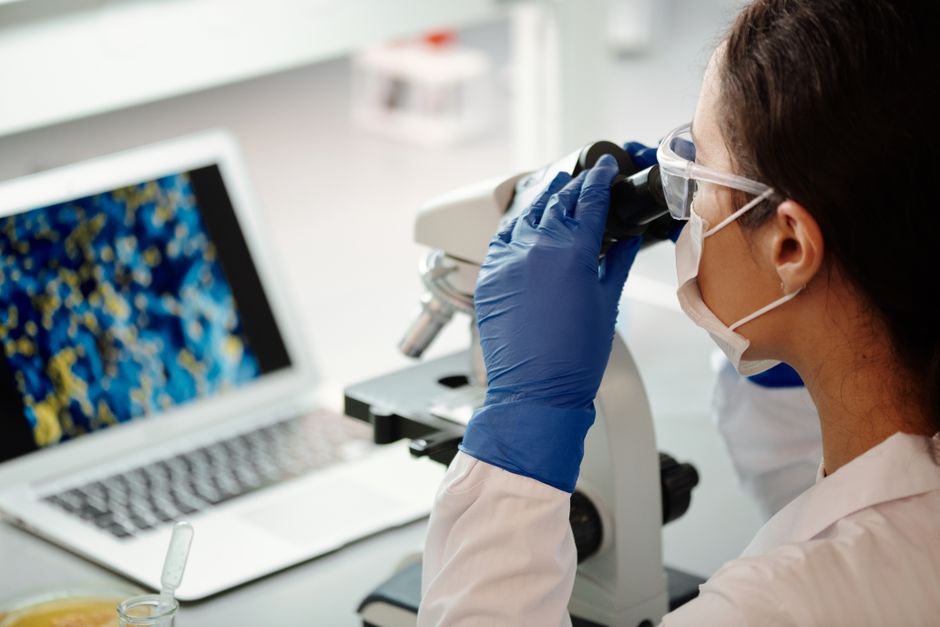 Scientist in lab coat examines samples using a microscope beside laptop, highlighting modern research techniques.