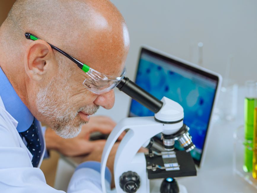 Senior scientist in lab coat using microscope, focused on research with laptop in background.